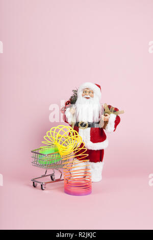 Ein Weihnachtsmann Figur Plüsch vor einem Einkaufswagen mit Regenbogen Feder im auf einem rosa Hintergrund. Farbharmonie. Minimale still life Stockfoto