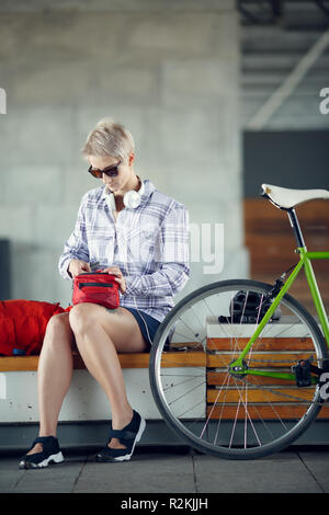 Foto des blonden Jungen in Kopfhörer sitzt auf der Bank neben dem grünen Fahrrad während des Tages Stockfoto