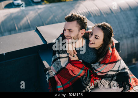 Hohe Betrachtungswinkel des jungen Paares im Plaid sitzen auf der Dachterrasse zusammen lachen Stockfoto