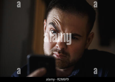 Der bärtige Mann mittleren Alters amazedly Suchen in dem dunklen Raum an das Smartphone. Close Up. Stockfoto