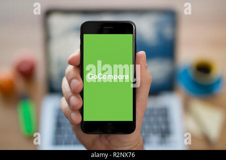 Ein Mann schaut auf seinem iPhone die zeigt die GoCompare logo, während saß an seinem Computer Schreibtisch (nur redaktionelle Nutzung). Stockfoto