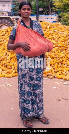 Angadihalli, Karnataka, Indien - November 2, 2013: Junge Mutter mit blauem Kleid hält schlafendes Baby in rot Schulter wickeln. Hintergrund ist Haufen gelb Stockfoto