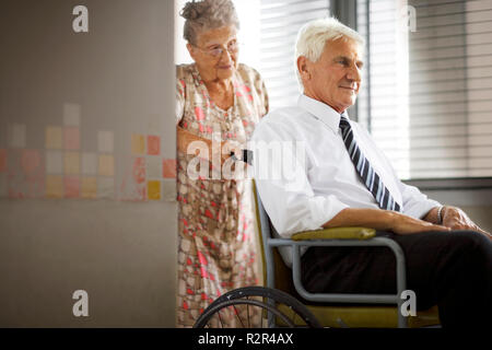 Eine ältere Frau drückt einen älteren Mann in einem Rollstuhl. Stockfoto