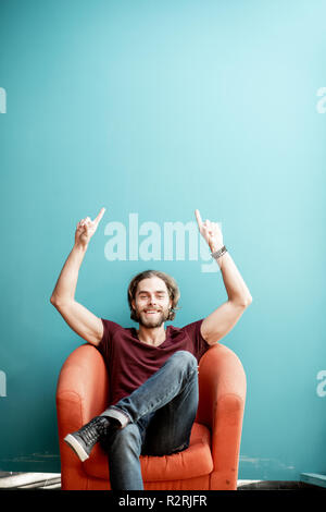 Portrait einer jungen kaukasischen bärtiger Mann mit langen Haaren, mit den Händen auf die bunten Hintergrund sitzen auf dem Stuhl. Bild mit Kopie Raum Stockfoto