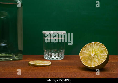 Eine halbe Limette mit unscharfen Kalk Schicht, Silver Tequila Shot Umrandet mit Salz und einer Flasche auf einen hölzernen Tisch im Landhausstil. Grüner Hintergrund. Stockfoto