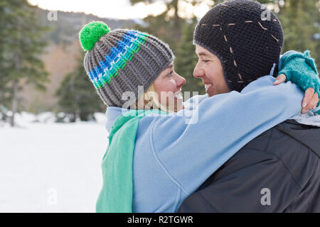 Glückliches Paar umarmen und sich gegenseitig an, während beide trugen gestrickte Hüte draußen im Schnee. Stockfoto