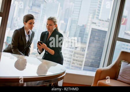 Mitte der erwachsenen Geschäftsfrau mit einem jüngeren Kollegen ein Mobiltelefon, während Sie an einem Tisch sitzen zusammen in einem Büro. Stockfoto