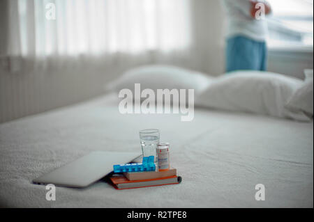 Geschlossen, Laptop, zwei Bücher, ein Glas Wasser, zwei Pille Flaschen und eine Medikation Dispenser sitzt auf einem Bett mit einer Person, die vor einem Fenster. Stockfoto