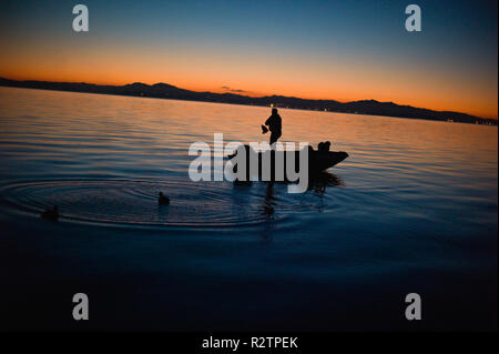 Der Mensch steht in der Mitte ein Boot und sammelt in entelockvögel mit seinem Hund am Bug beim Schwimmen in einem See, wie die Sonne. Stockfoto