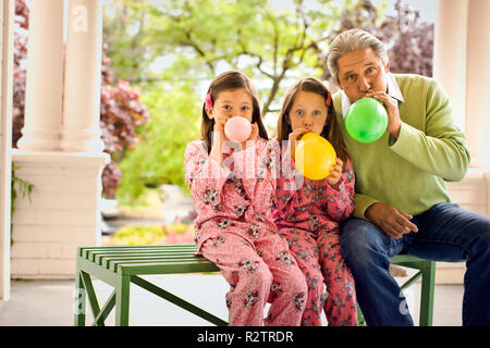 Zwei junge Mädchen in passenden Schlafanzug sitzen mit einem reifen Mann auf einer Bank auf einer Veranda und ein Ballon jeweils als sie posieren für ein Portrait. Stockfoto