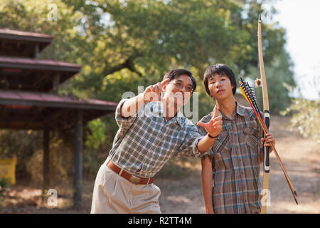 Lächelnd Mitte der erwachsene Mann Lehre sein halbwüchsiger Sohn wie ein Pfeil und Bogen zu benutzen. Stockfoto