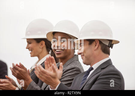 Drei lachenden Kollegen in Anzügen und hardhats applaudieren nach dem Spatenstich für ein neues Gebäude. Stockfoto
