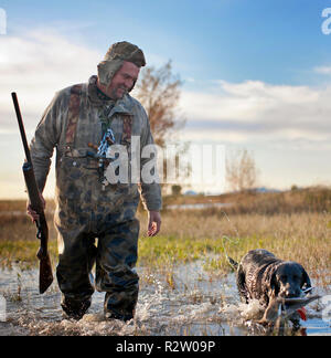Duck Hunter zu Fuß durch ein flacher See neben seinem Hund, hält eine tote Ente in den Mund. Stockfoto