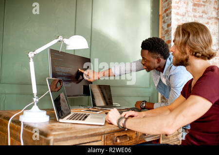 Zwei junge Programmierer mit Programmcode am Arbeitsplatz sitzen mit drei Monitoren im Büro auf die Grüne Wand Hintergrund Stockfoto