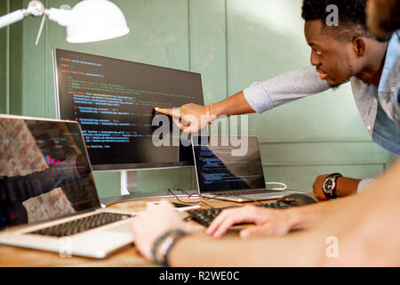 Zwei junge Programmierer mit Programmcode am Arbeitsplatz sitzen mit drei Monitoren im Büro auf die Grüne Wand Hintergrund Stockfoto