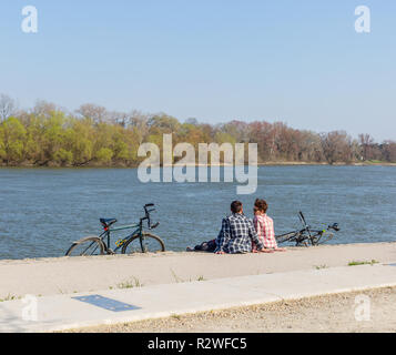 Szentendre, Ungarn - April 08, 2018: Romantische junge Paar in Karohemden sitzt am Ufer des Flusses mit Fahrrädern. Active Living Konzept. Platz für Stockfoto