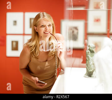 Positive Frau mit Brille und Smartphone Besuch von historischen Museum und Vitrinen mit Exponaten auf der Suche Stockfoto