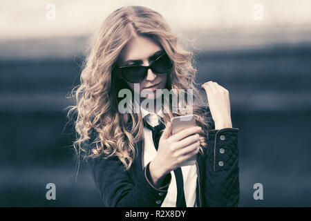 Blond business Frau mit Handy in der city street Stilvolle Mode Modell mit Langen lockigen Haare mit schwarzen Jacke und Sonnenbrille Stockfoto