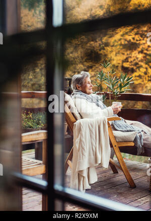 Eine ältere Frau sitzt im Freien auf der Terrasse im Freien an einem sonnigen Tag im Herbst. Stockfoto