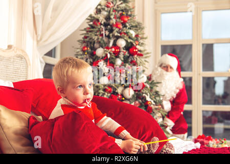 Wenig traurig Junge sitzt auf einem Bett in einem festlichen Schlafzimmer. Die Real Santa Claus ist das Verstecken von ihm hinter dem Weihnachtsbaum. Stockfoto