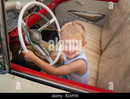 Schönes Kind sitzt auf alten amerikanischen Autos der 50er/60er Jahre, hält Lenkrad und tut so, als ob es zu fahren. Kaukasische weiß baby boy. Stockfoto