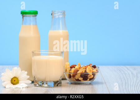 Zwei Flaschen und ein Glas frische Milch, Glasschale mit Müsli und Kamille Blüte auf blauem Hintergrund Stockfoto