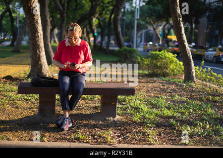 Reifen Taiwanese chinesische Frau sitzen auf einer Bank in einem städtischen Park an Ihrem Mobiltelefon, die in der Abendsonne. Stockfoto