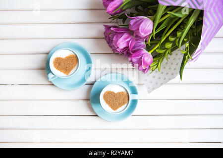 Zwei Tassen Cappuccino mit einem Herzförmigen Symbol und lila Tulpen auf einer hölzernen Hintergrund. Stockfoto