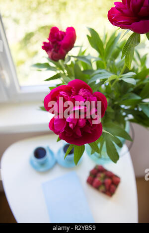 Erdbeere, Buch, Blumenstrauß aus schönen Pfingstrosen und zwei Tassen Kaffee auf weißer Tisch. Stockfoto