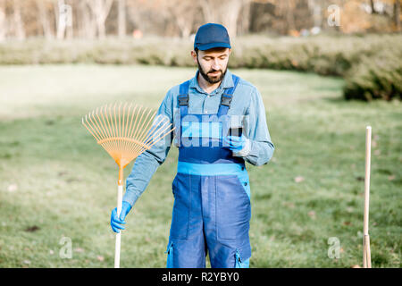 Professionelle sweeper in Uniform mit Phone, der während der Arbeit im Garten Stockfoto