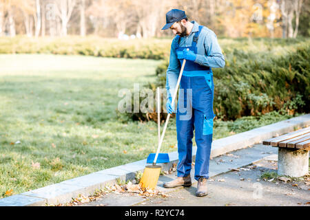 Professionelle sweeper in Uniform fegt Blätter mit Besen und Schaufel auf der Straße Stockfoto