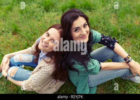 Smiling brunette und rötlich Mädchen zurück sitzen im Gras zurück und Blick in die Kamera, Beine leicht gebeugt in die Knie. Top Winkel anzeigen Stockfoto
