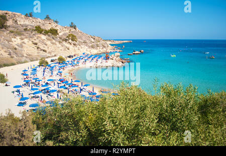 Bild von Konnos Beach in der Nähe von Agia Napa, Zypern. Viele blaue Liegestühle und Sonnenschirme auf weißem Sand in der Nähe transparent blau Wasser in einer Bucht und felsigen Hügeln. Stockfoto