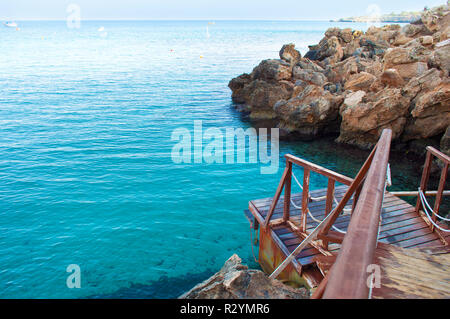 Bild eines Holz- braun Pier mit Seilen in der Nähe von fantastischen blauen Meer Wasser in der Nähe von Felsen auf dem Hintergrund der vielen Boote an einem wolkenlosen Tag fallen. Zypern, Konnos Stockfoto