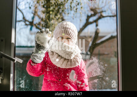 Sieben Jahre alten süße Mädchen im Winter Kleidung steht vor der Tür, auf der Straße mit Schnee in den Händen und Suchen in das Haus ein Lächeln auf den Lippen. Stockfoto