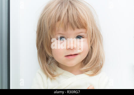 Headshot von adorable cute Blonde weibliches Kind auf Kamera mit schweren umgekippt Gesichtsausdruck. Grinsen auf dem Gesicht. Hübsches kleines Mädchen mit kaukasischen Merkmale und Kratzer auf der Nase. Attraktive baby Stockfoto