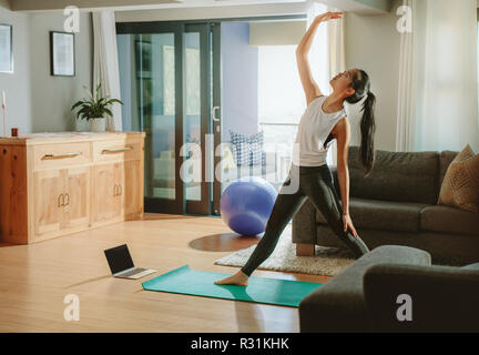 Frau, die in ihrem Wohnzimmer. Frau stehend auf Yoga Matte und Stretching mit Laptop auf dem Boden. Stockfoto