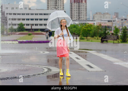 Junge hübsche Mädchen mit zwei Litzen in den gelben Stiefeln und mit transparentem Dach steht in der Nähe von Brunnen. Regnerischen Tag in der Stadt. Stockfoto