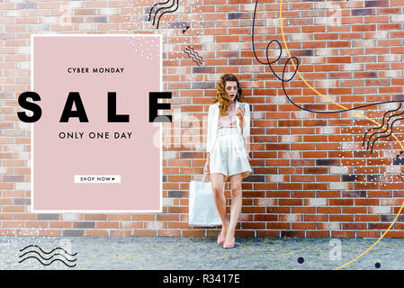 Schockiert junge Frau mit Einkaufstüten Sie Ihr Smartphone vor der Mauer, Cyber Monday banner Stockfoto