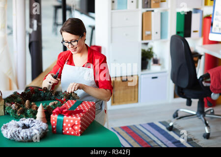 Kreative junge Frau in Arbeitskleidung steht die Tabelle mit Xmas-Personal und beenden mit dekorativen Kranz Stockfoto