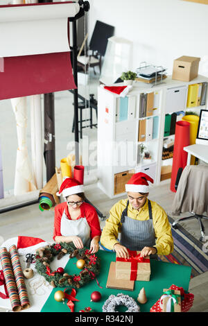 Junge kreative Mann und Frau in Santa Caps, pre-xmas Arbeiten in Design Studio Stockfoto