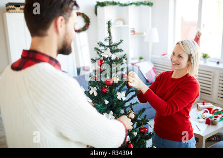 Glückliche junge Frau auf der Suche nach ihrem Mann, während beide schmücken Weihnachtsbaum zu Hause Stockfoto