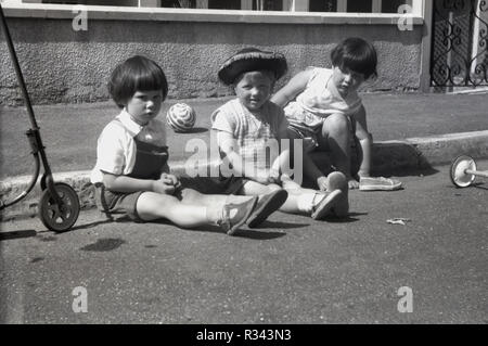 1960, historische, drei kleine Kinder draußen auf der Straße sitzend durch das Pflaster, mit Ihrer Kugel und Motorroller, England, UK. Der Junge trägt eine mexikanische Cowboyhut. Stockfoto