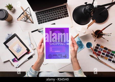 7/8-Ansicht von Designer holding Tablet mit Shopping App auf dem Bildschirm im Büro Schreibtisch mit Künstlerbedarf, flach Stockfoto