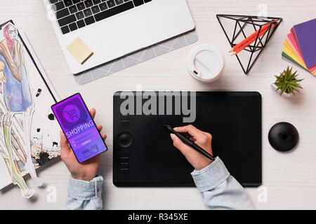 7/8-Ansicht von Designer mit Grafiktablett, Pen und Smartphone mit Shopping App auf dem Bildschirm, flach Stockfoto