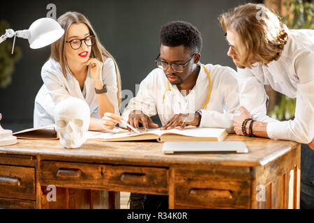 Gruppe von einem Jungen multi Ethnizität Ärzte oder Medizinstudenten in Uniform arbeiten mit Buch und Zeichnungen auf dem Tisch im Büro oder Klassenzimmer Stockfoto