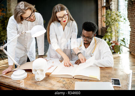 Gruppe von einem Jungen multi Ethnizität Ärzte oder Medizinstudenten in Uniform arbeiten mit Buch und Zeichnungen auf dem Tisch in der retro Office Interior Stockfoto