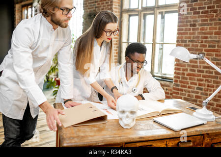 Gruppe von einem Jungen multi Ethnizität Ärzte oder Medizinstudenten in Uniform arbeiten mit Buch und Zeichnungen auf dem Tisch im Büro oder Klassenzimmer Stockfoto