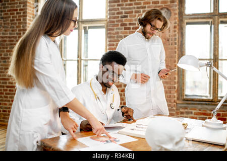 Gruppe von einem Jungen multi Ethnizität Ärzte oder Medizinstudenten in Uniform arbeiten mit Buch und Zeichnungen auf dem Tisch im Büro oder Klassenzimmer Stockfoto