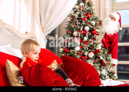 Wenig traurig Junge sitzt auf einem Bett in einem festlichen Schlafzimmer. Die Real Santa Claus ist das Verstecken von ihm hinter dem Weihnachtsbaum. Stockfoto